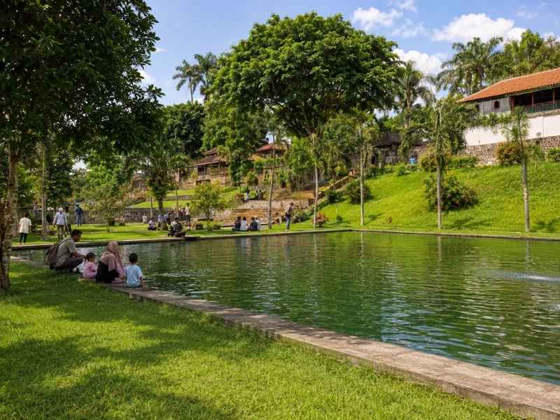 Keluarga bersantai di tepi kolam Taman Narmada yang dikelilingi pepohonan hijau